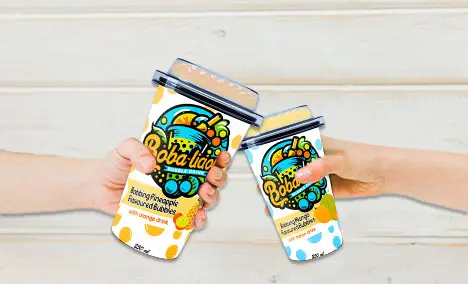 Two hands toasting with Bobalicious Pineapple and Mango Bubble Tea 250ml cups against a light wooden background.