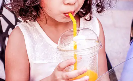 Young girl sipping an orange drink through a straw in a clear plastic cup.