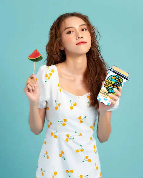 Young woman in a cherry-print dress holding a Bobalicious Mango Bubble Tea and a slice of watermelon against a blue background.